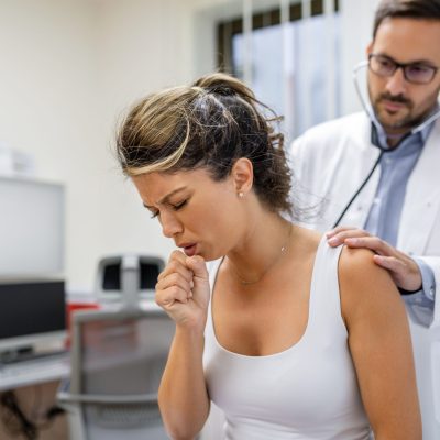 Young female patient in the clinic suffered from pneumonia, she is coughing the doctor listens to the wheezing in the lungs with a stethoscope.